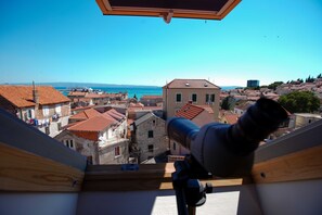 A telescope on a balcony overlooking a coastal town with red-roofed buildings and a clear blue sky.