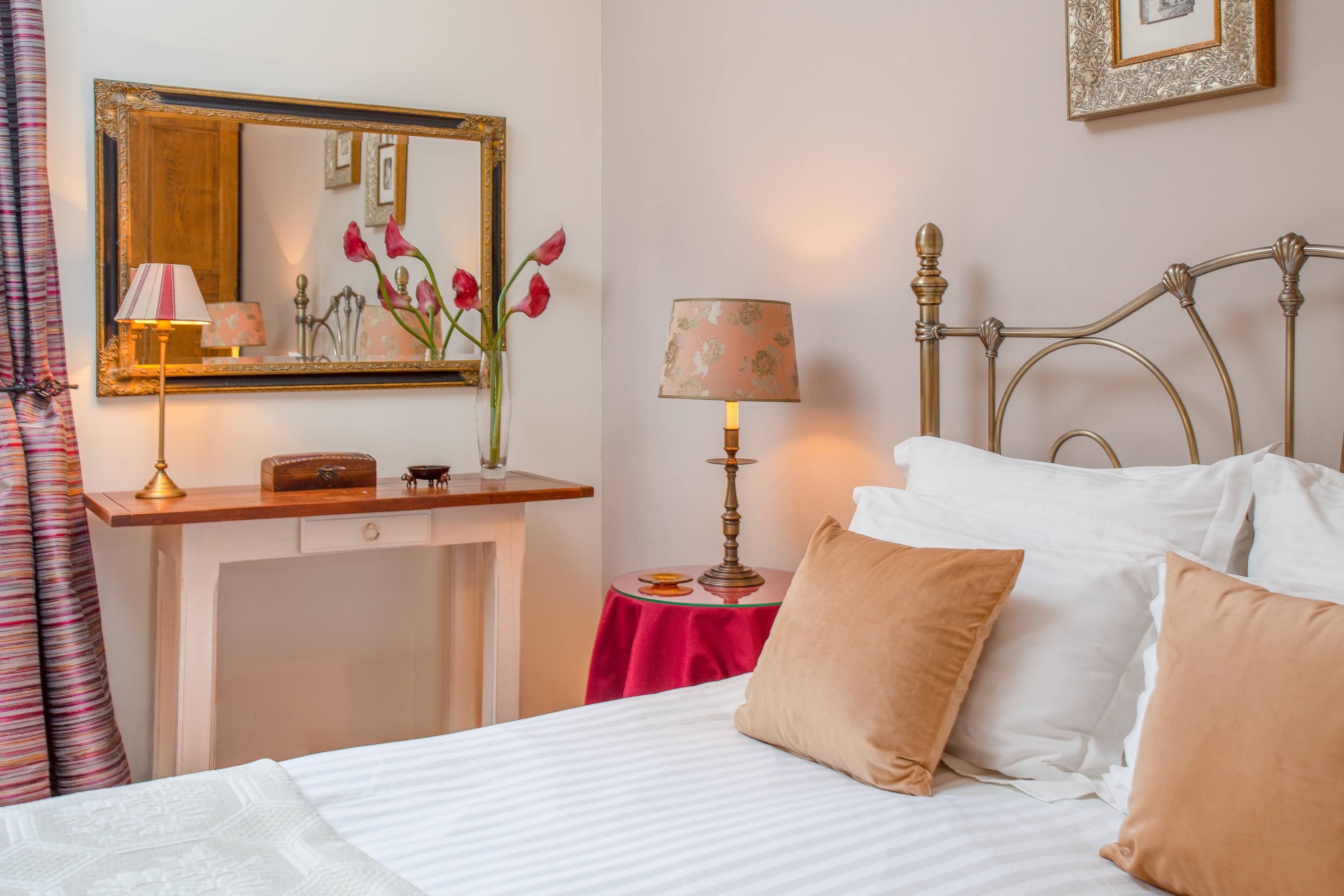 A hotel room with a bed, a bedside table with a lamp, a mirror, and a small table with a lamp.
