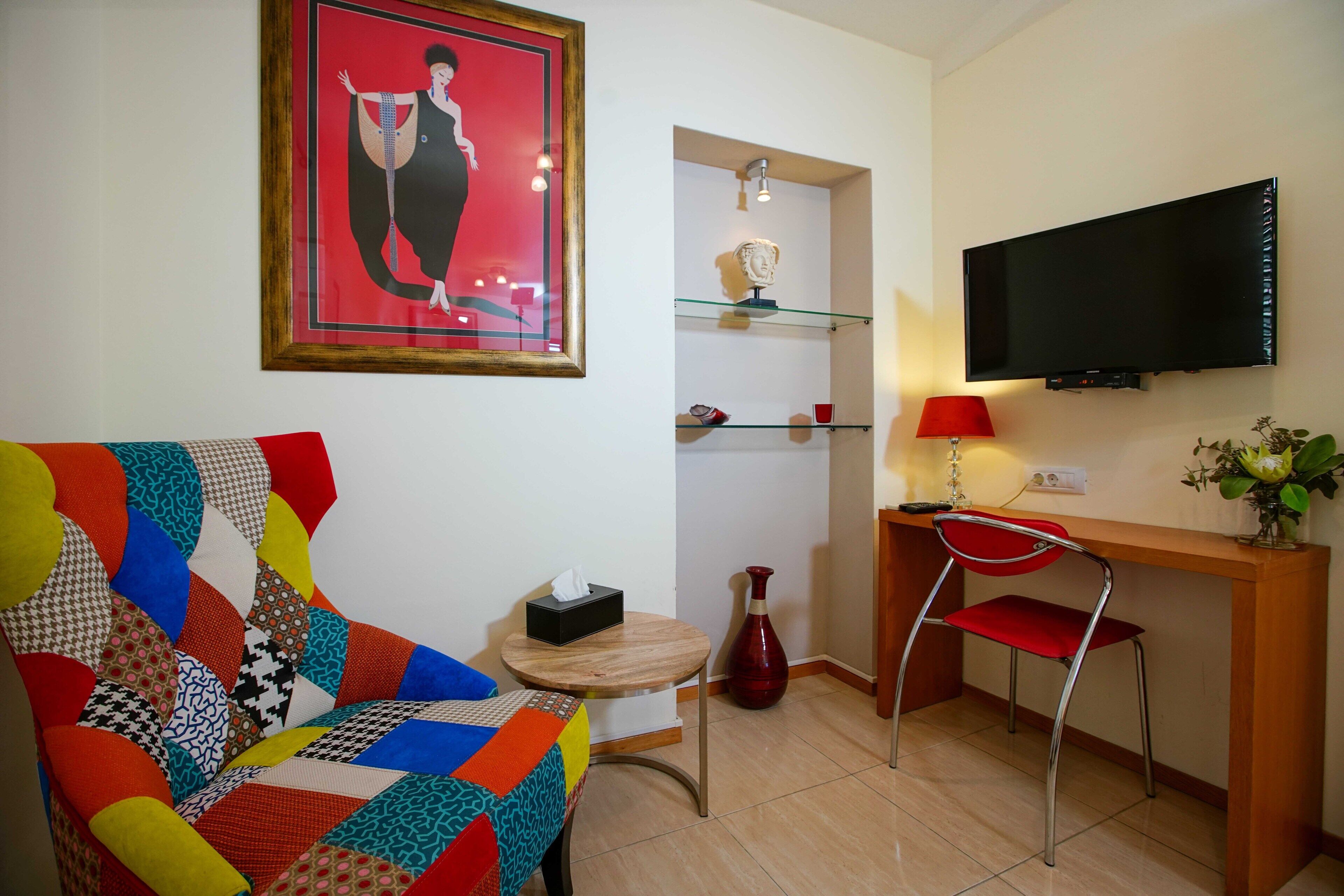 Un salon avec un canapé aux couleurs vives, une petite table ronde sur laquelle se trouve une boîte à mouchoirs, un vase rouge, un fauteuil rouge, un téléviseur fixé au mur et une étagère ornée d’objets décoratifs.
