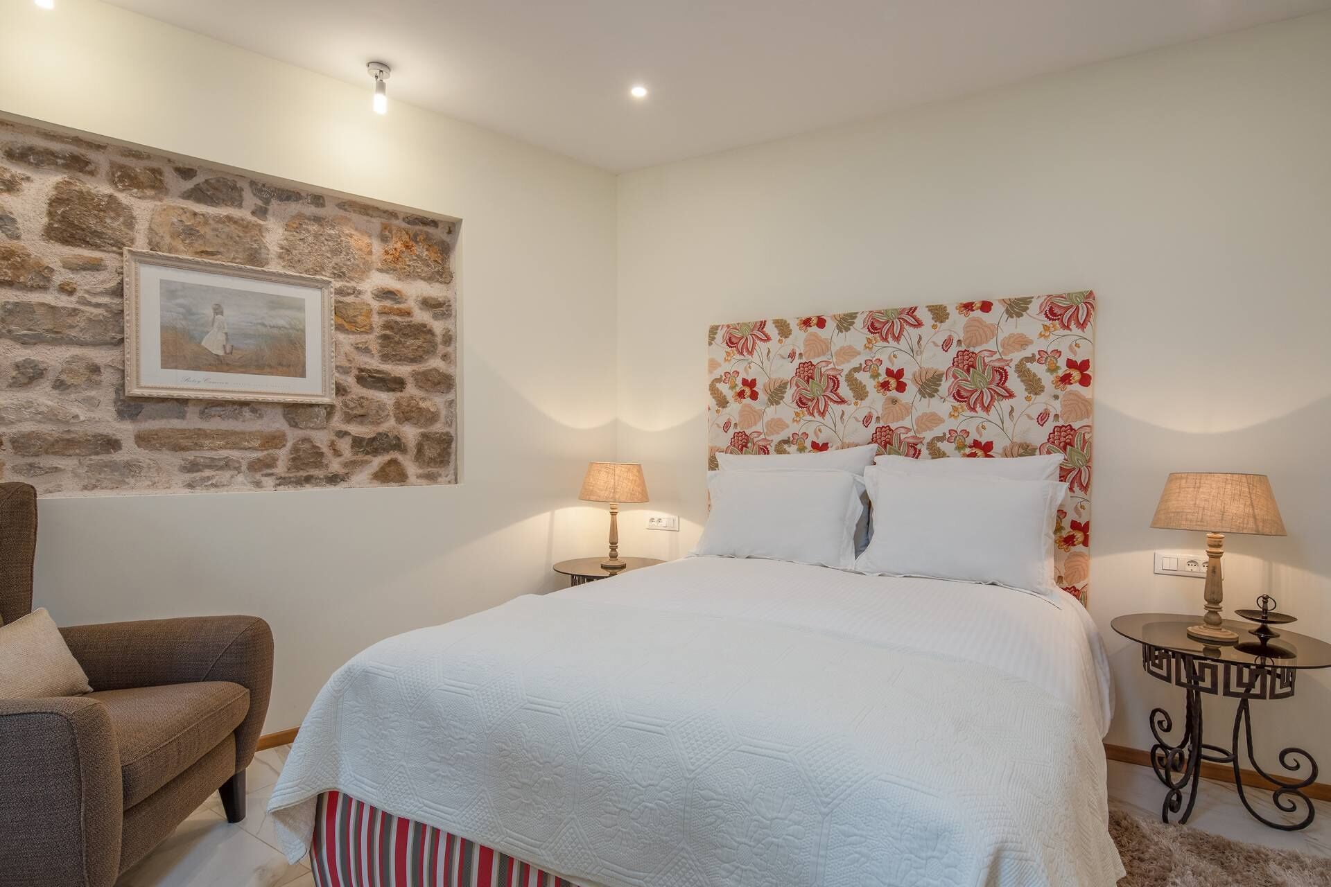 A bedroom with a stone wall, a bed with floral headboard, two bedside tables with lamps, and a chair.