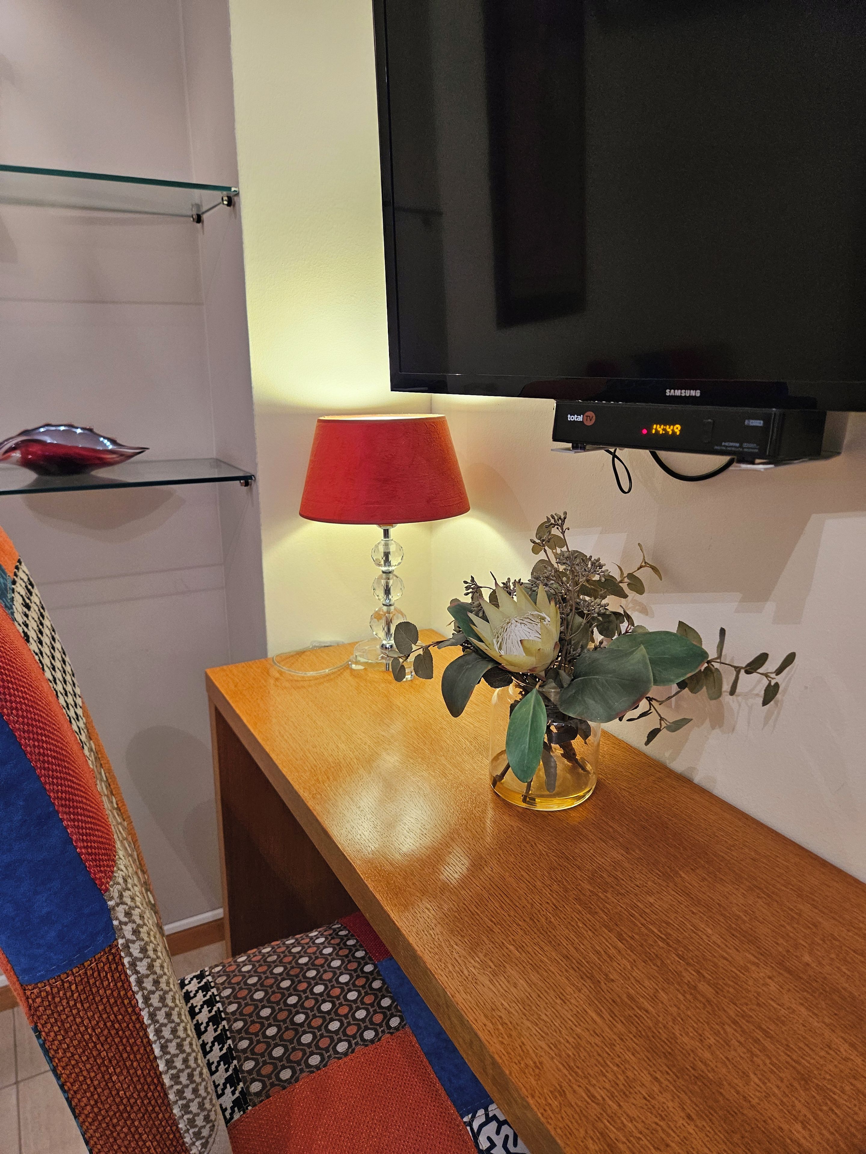 Een kamer met een houten tafel, een rode lamp, een bloevas, een televisie en een plank met een decoratieve schaal.