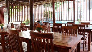 A restaurant with wooden tables and chairs, a thatched roof, and large windows. - Duy An Guest House (Mui Ne)
