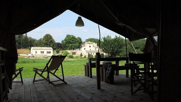 An outdoor wooden deck with two folding chairs and a table, overlooking a grassy area with buildings in the background. - Les Ânes de Forcalquier (Forcalquier)