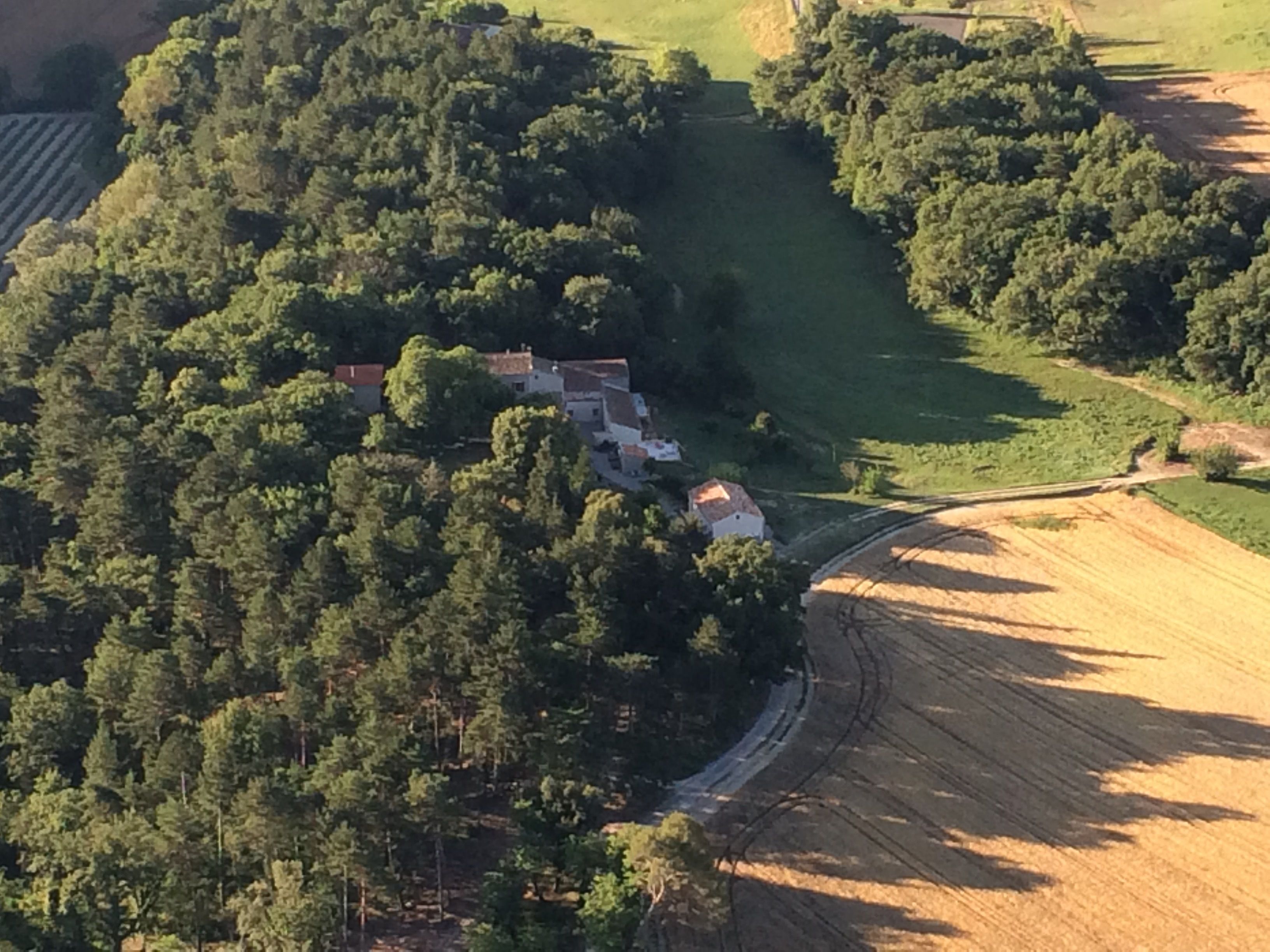 Vista aérea de uma área rural com um conjunto de casas rodeadas por floresta densa. Uma estrada sinuosa serpenteia pela paisagem, conduzindo a campos abertos.