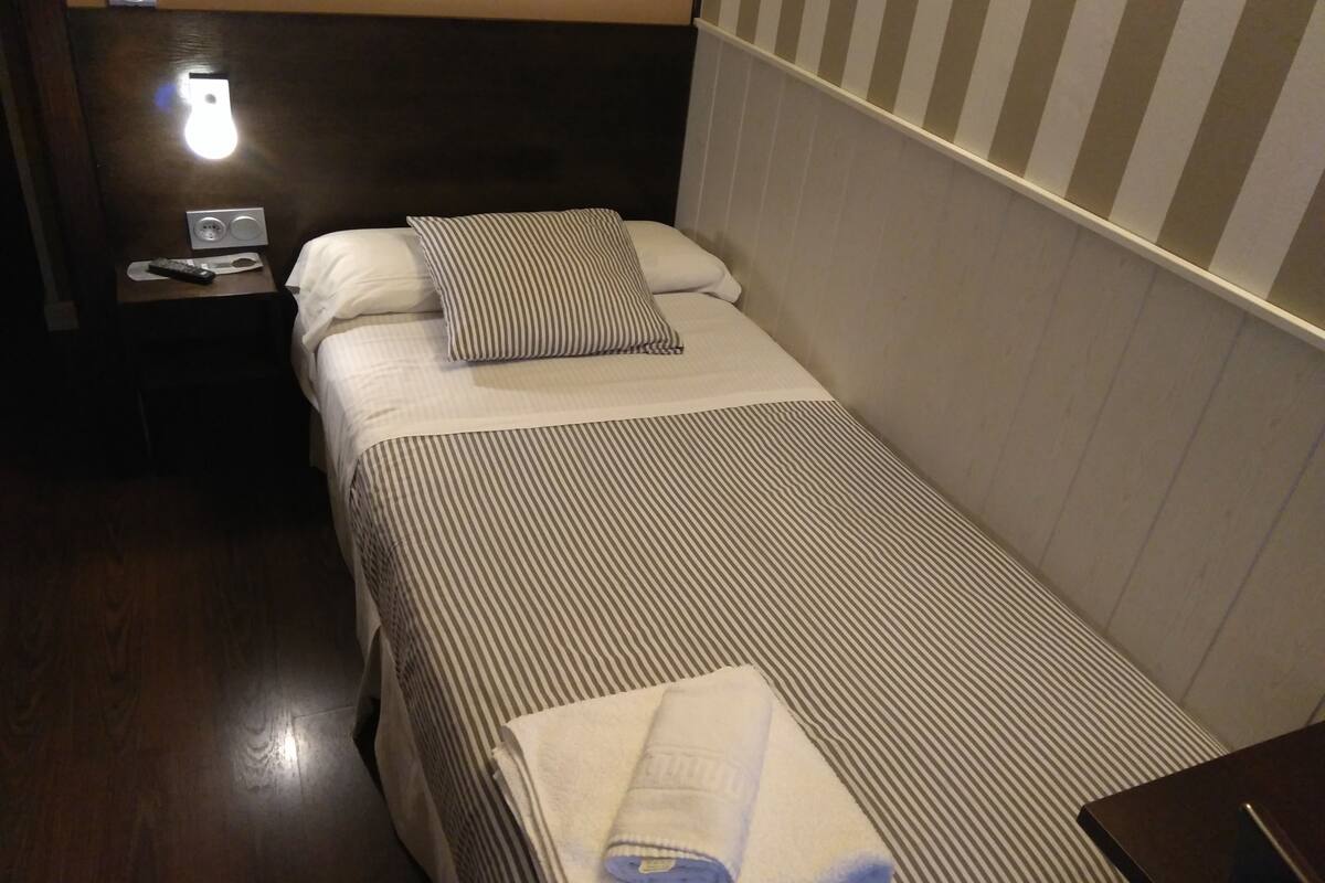 Un lit simple avec une literie à rayures, un oreiller, une table de chevet avec une télécommande et une lampe fixée au mur.