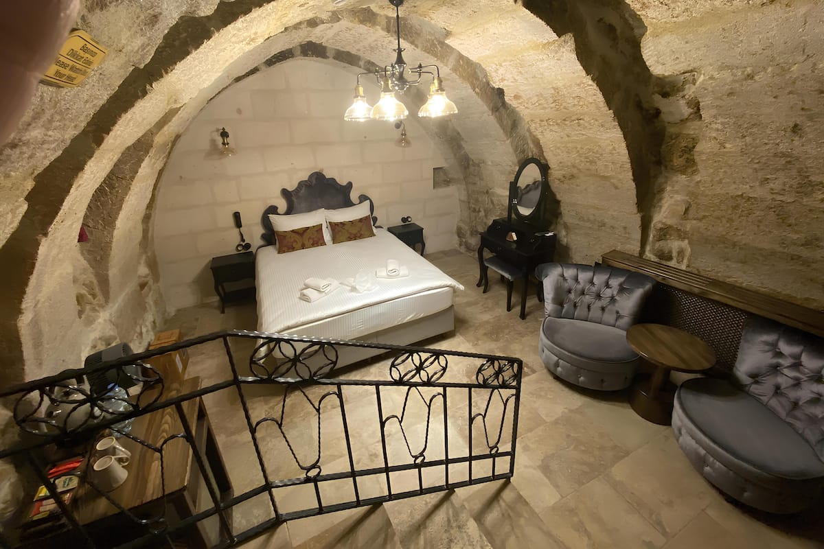 A bedroom with a stone arched ceiling, a bed with white linens, a vanity with a mirror, and a seating area with a cushioned chair and ottoman.