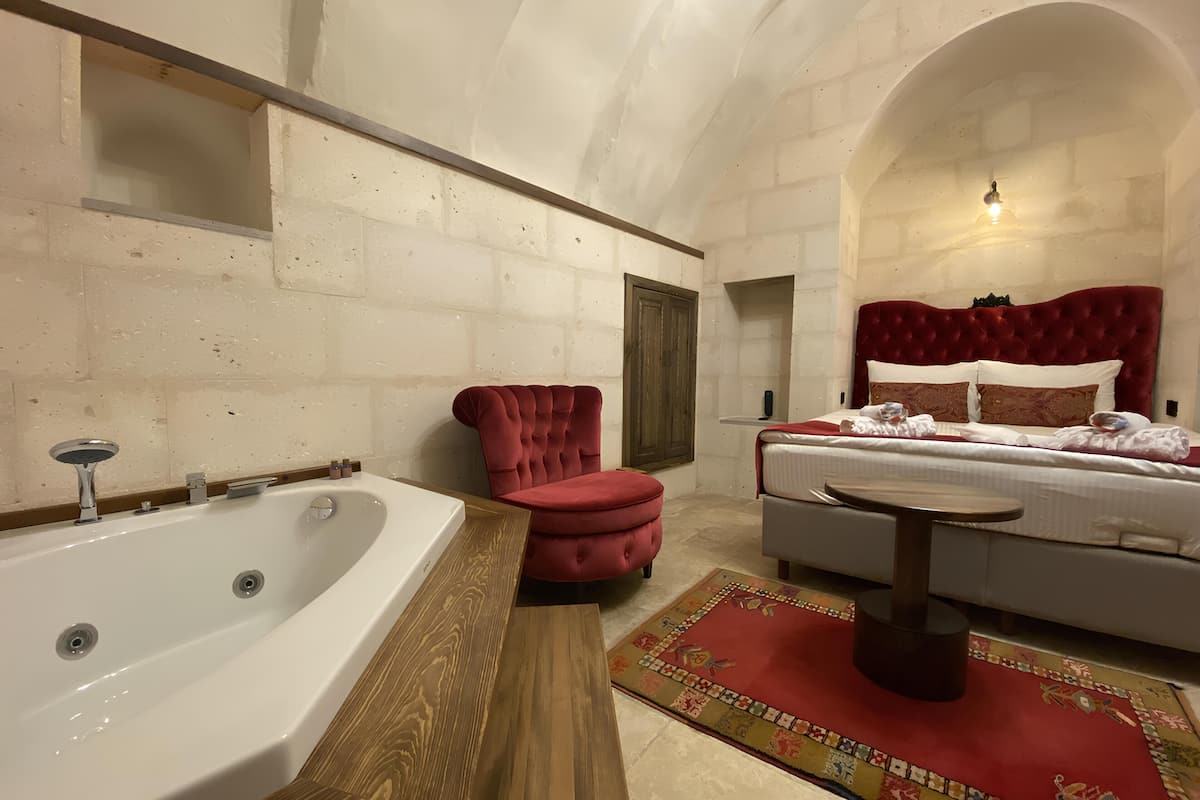 A bedroom with a large bathtub, a red armchair, a bed with a red headboard, and a wooden side table.