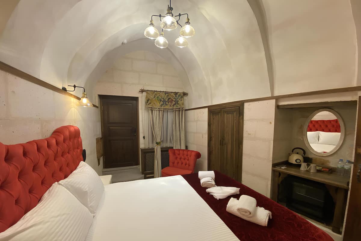 A hotel room with a red upholstered headboard, a large bed, a red velvet bedspread, a round mirror, a small table with a kettle, and a window with a view of the outside.