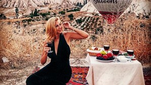 A woman in a black dress sitting on a patterned rug outdoors with a table set for a meal, surrounded by a scenic landscape with rock formations and hot air balloons in the background. - Sevdali Konaklar (Avanos)
