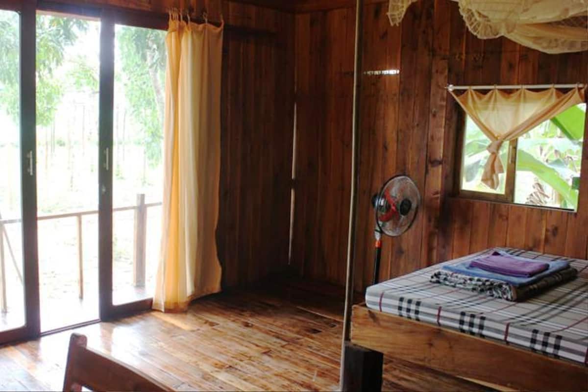 A wooden room with a bed, a chair, a fan, and a window with curtains.