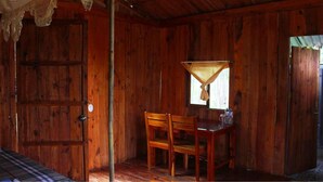 Una habitación de madera con una sola silla de madera y una mesa, una ventana con cortina, y una puerta con manija.