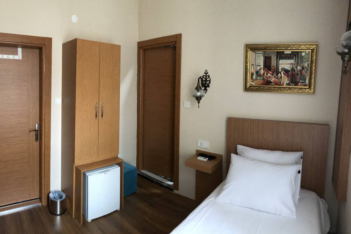 A hotel room with a bed, a small refrigerator, wooden wardrobes, a wall-mounted lamp, and a framed painting.