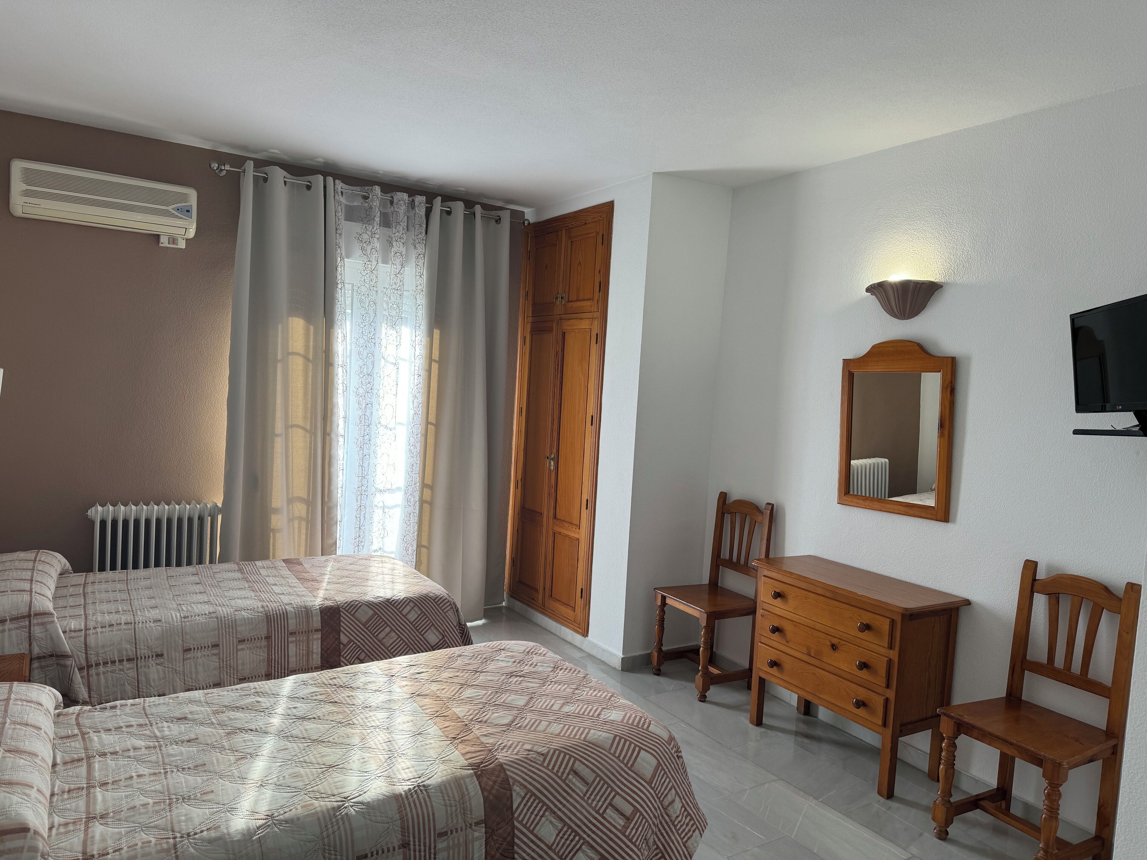 A hotel room with two beds, a wooden dresser, a desk, a chair, a radiator, a TV, and a mirror on the wall.