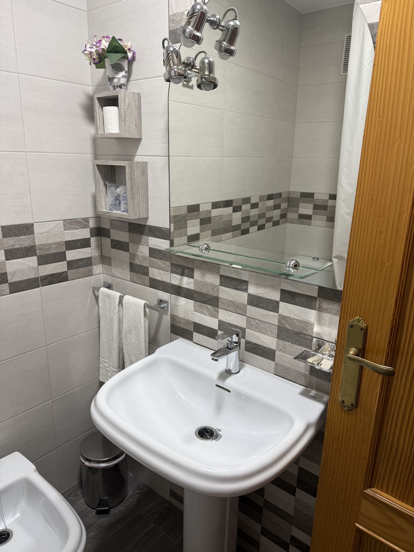 A bathroom with a white sink, a bidet, a glass shower enclosure, and a tiled wall with a checkered pattern.
