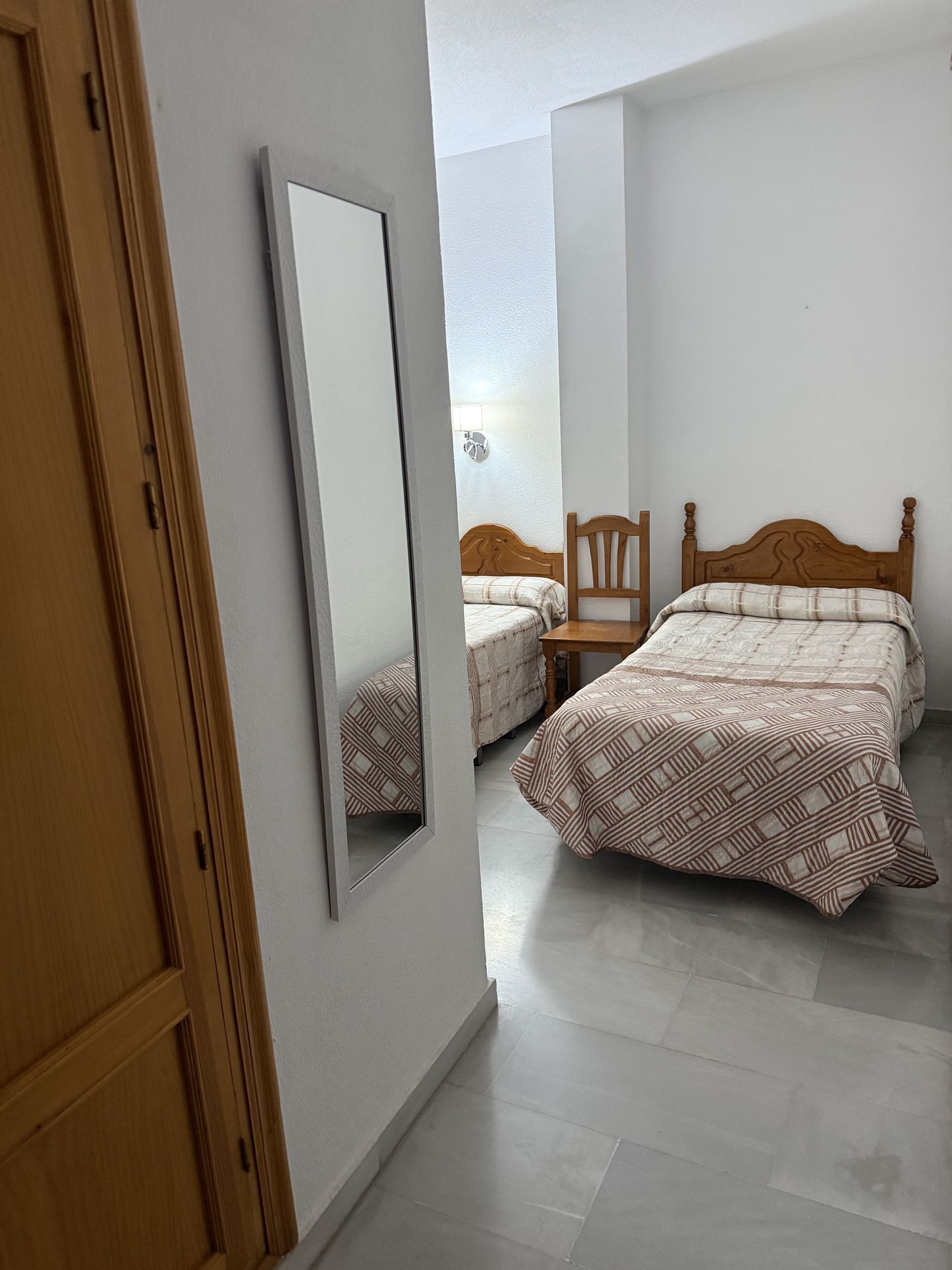 A bedroom with a wooden wardrobe, a bed with a patterned bedspread, a chair, and a bedside table.