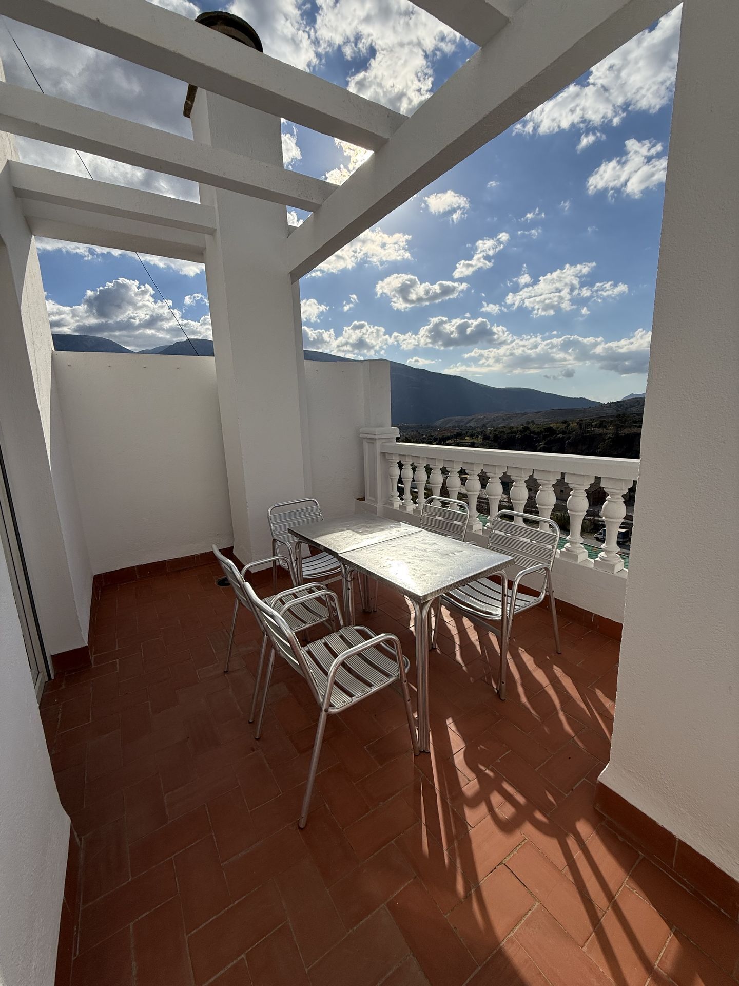 A balcony with a white railing, a table with four chairs, and a view of distant mountains and clouds.