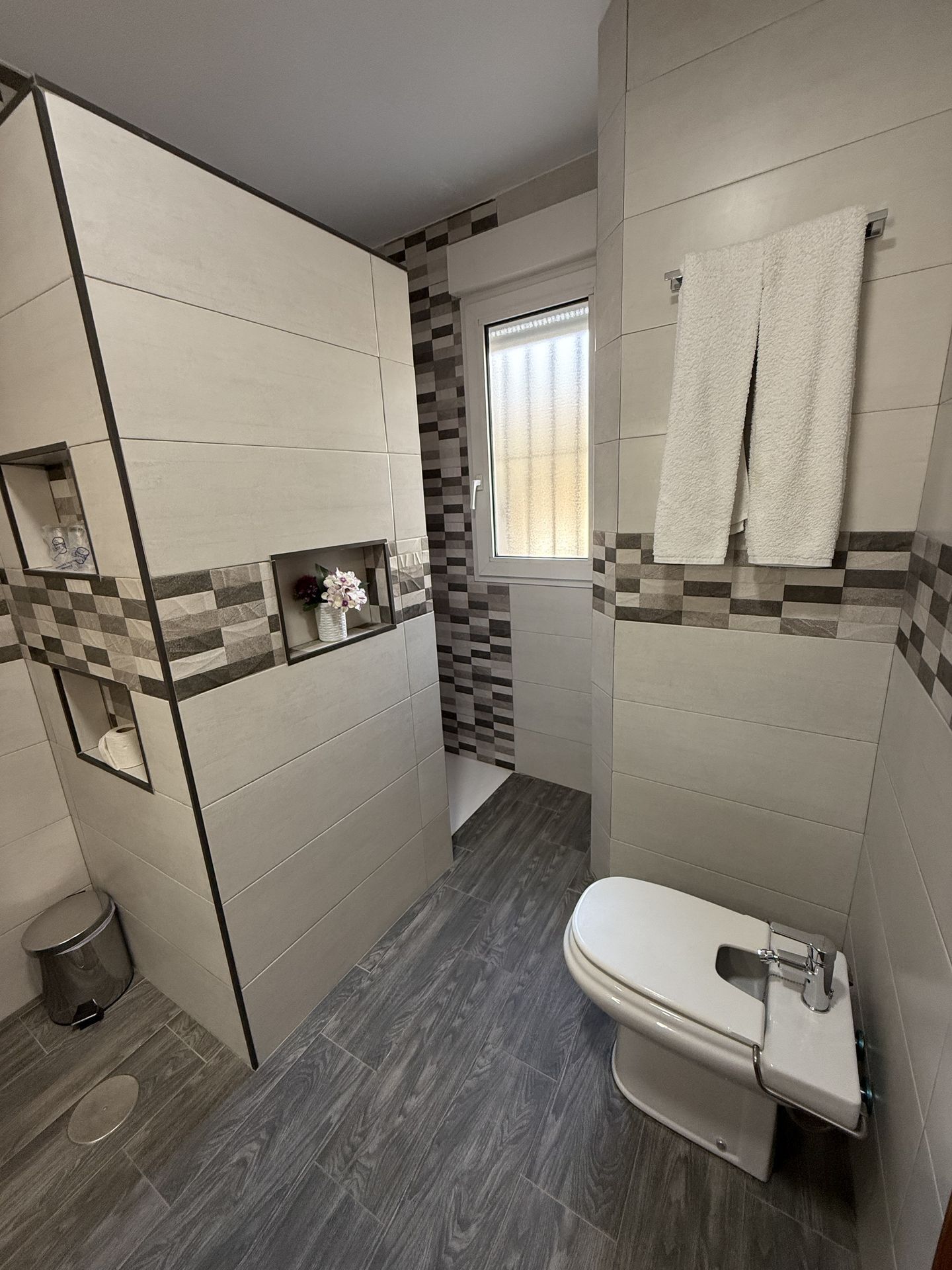 Un baño moderno con inodoro, lavamanos y espejo. Hay una ventana con vistas al exterior y las paredes presentan un diseño de cuadros.