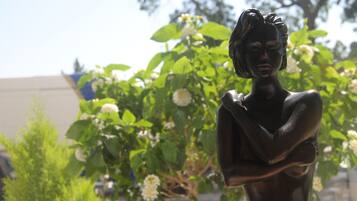 A statue of a woman with her arms crossed, set against a backdrop of green foliage and white flowers.