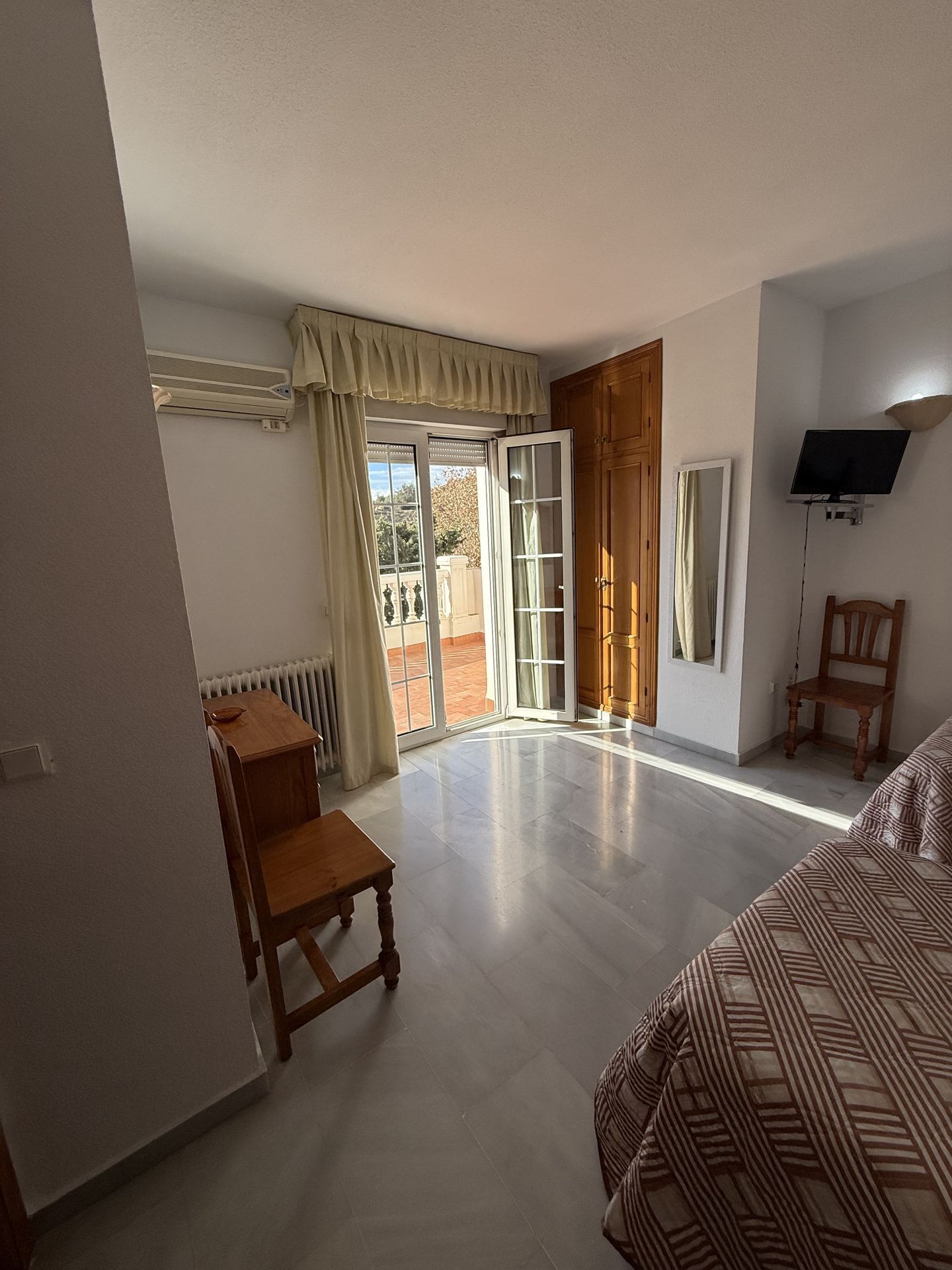 A room with a bed, a wooden chair, a small table, a radiator, a TV, and a sliding glass door leading to an outdoor area.
