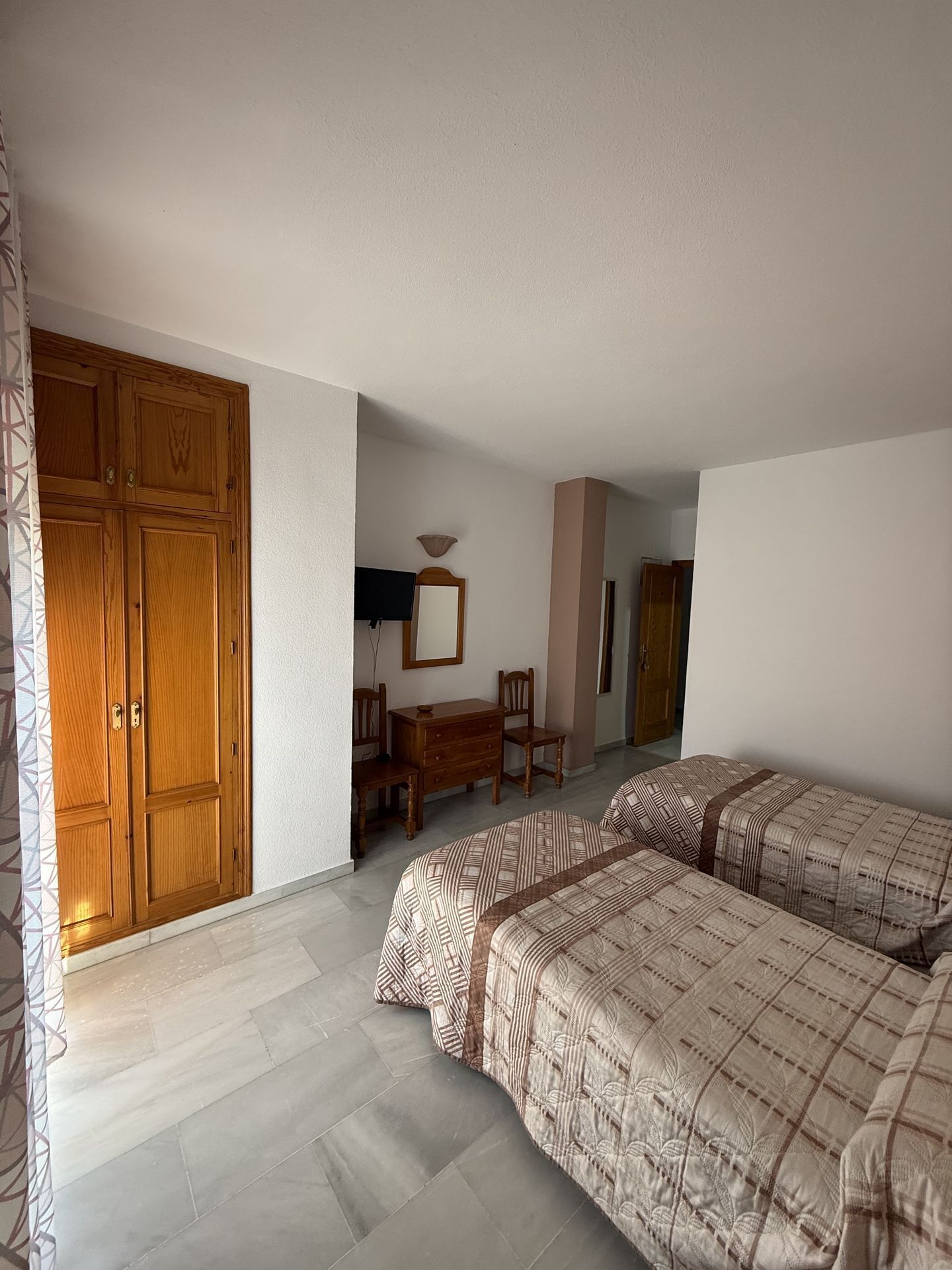 A bedroom with two beds, a wooden wardrobe, a desk with a chair, and a mirror on the wall.