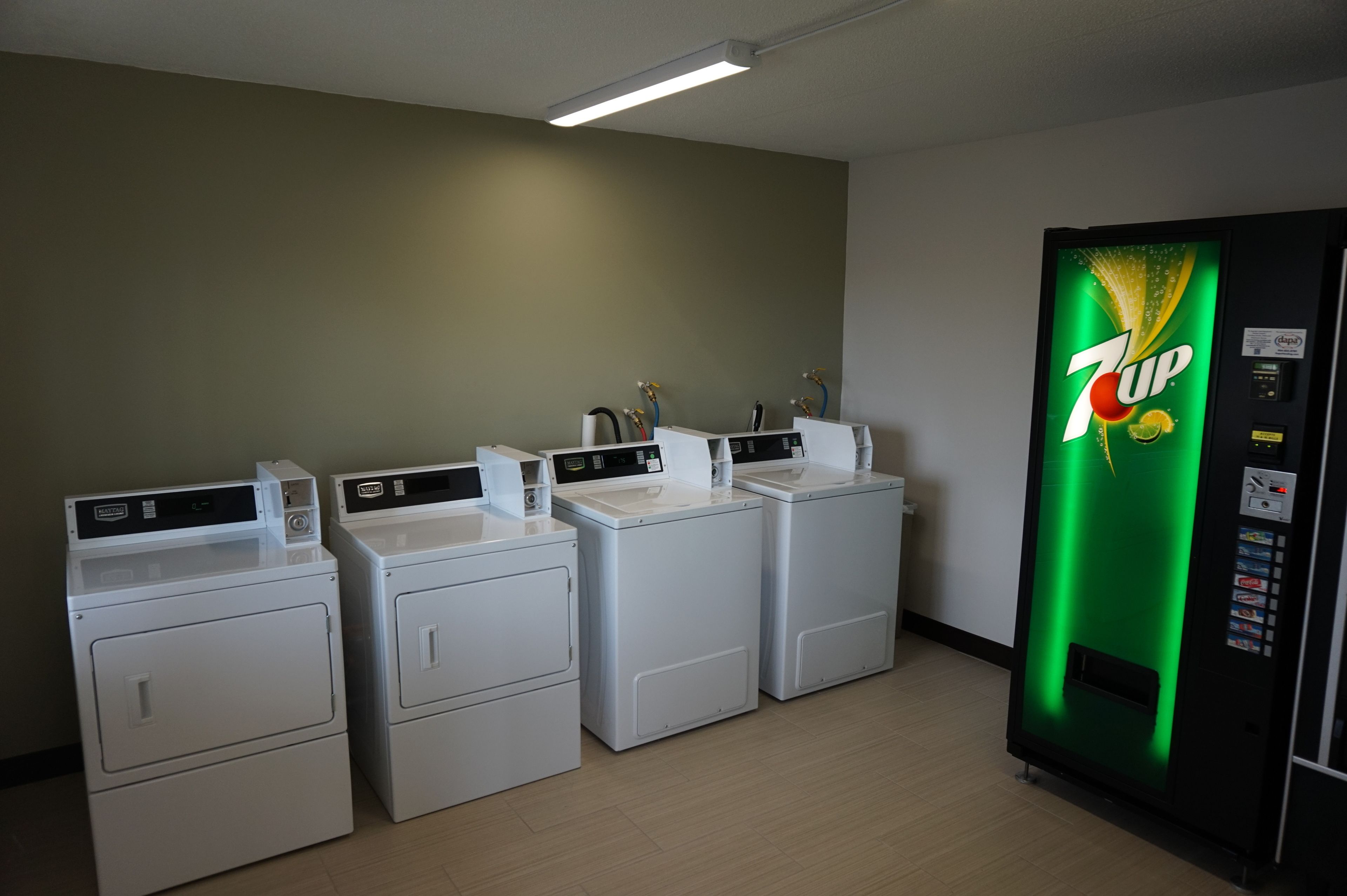 A laundry area with four white washers and dryers, a 7UP vending machine, and a green wall.