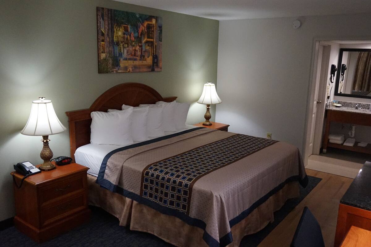 A hotel room with a bed, two bedside lamps, a nightstand with a phone, a painting on the wall, and a bathroom visible through an open door.