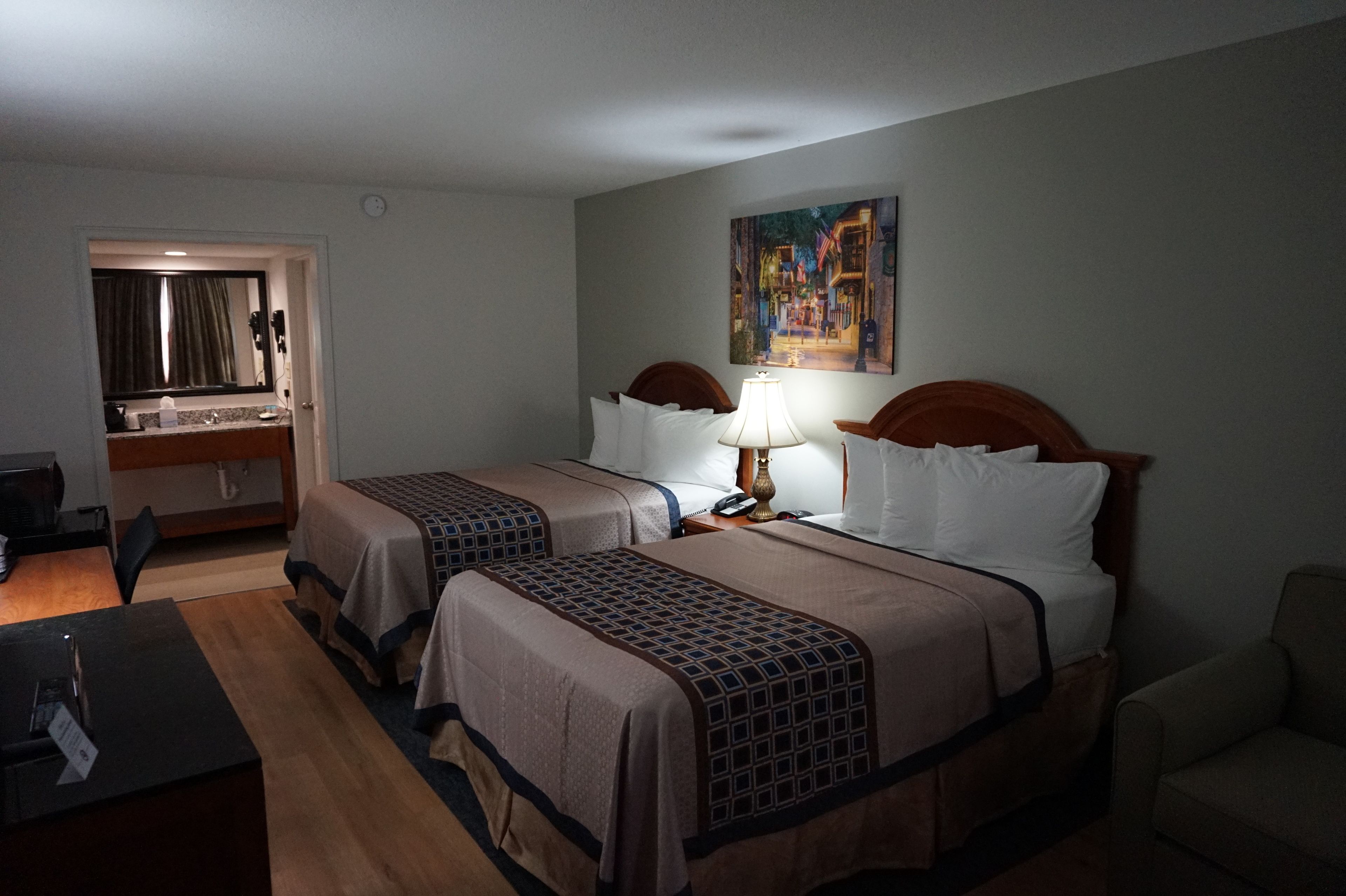 A hotel room with two beds, a desk, a chair, a painting on the wall, and a bathroom visible through an open door.