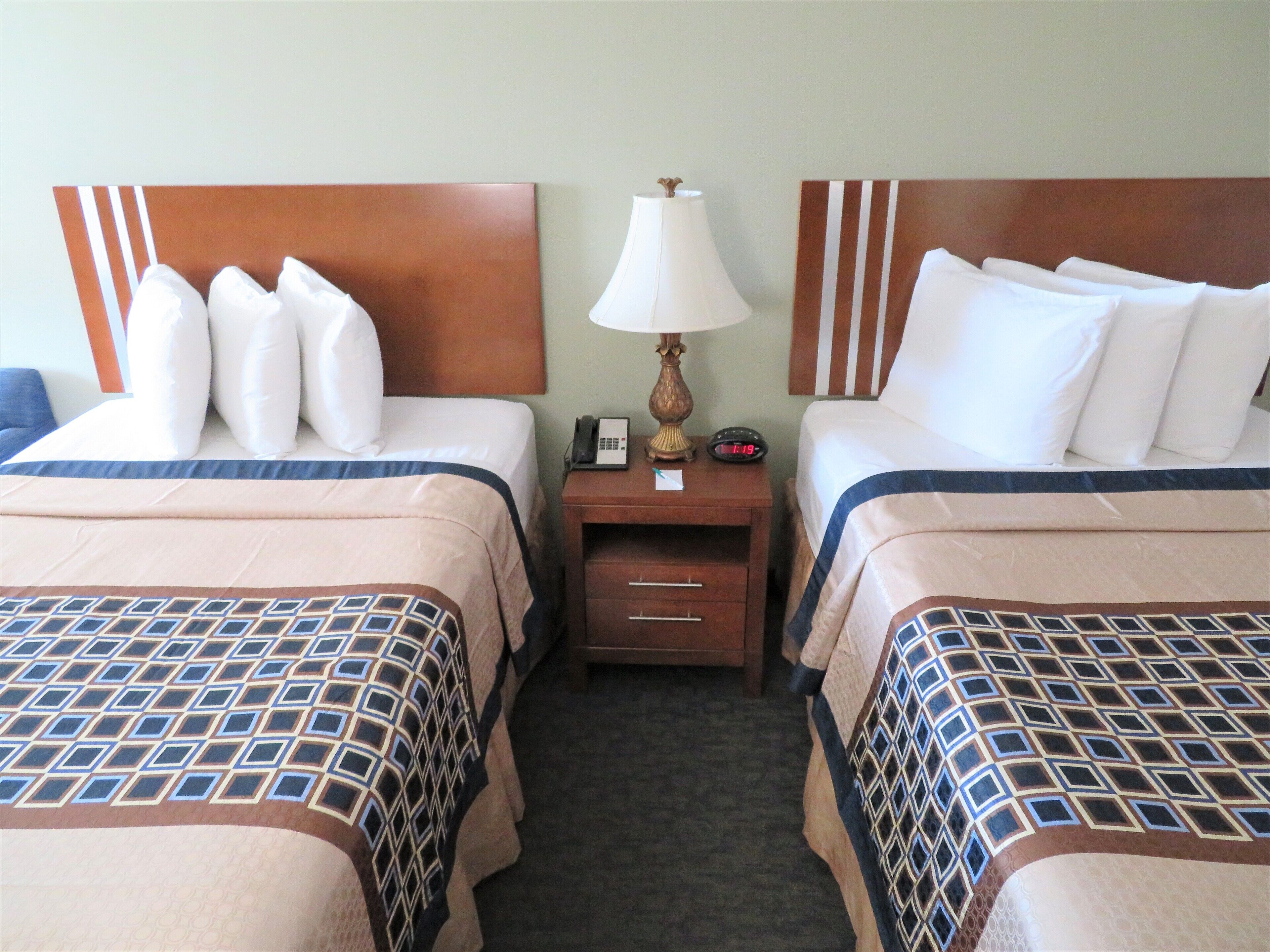 A hotel room with two beds, each with a headboard, a bedside table with a lamp, a clock, and a drawer.
