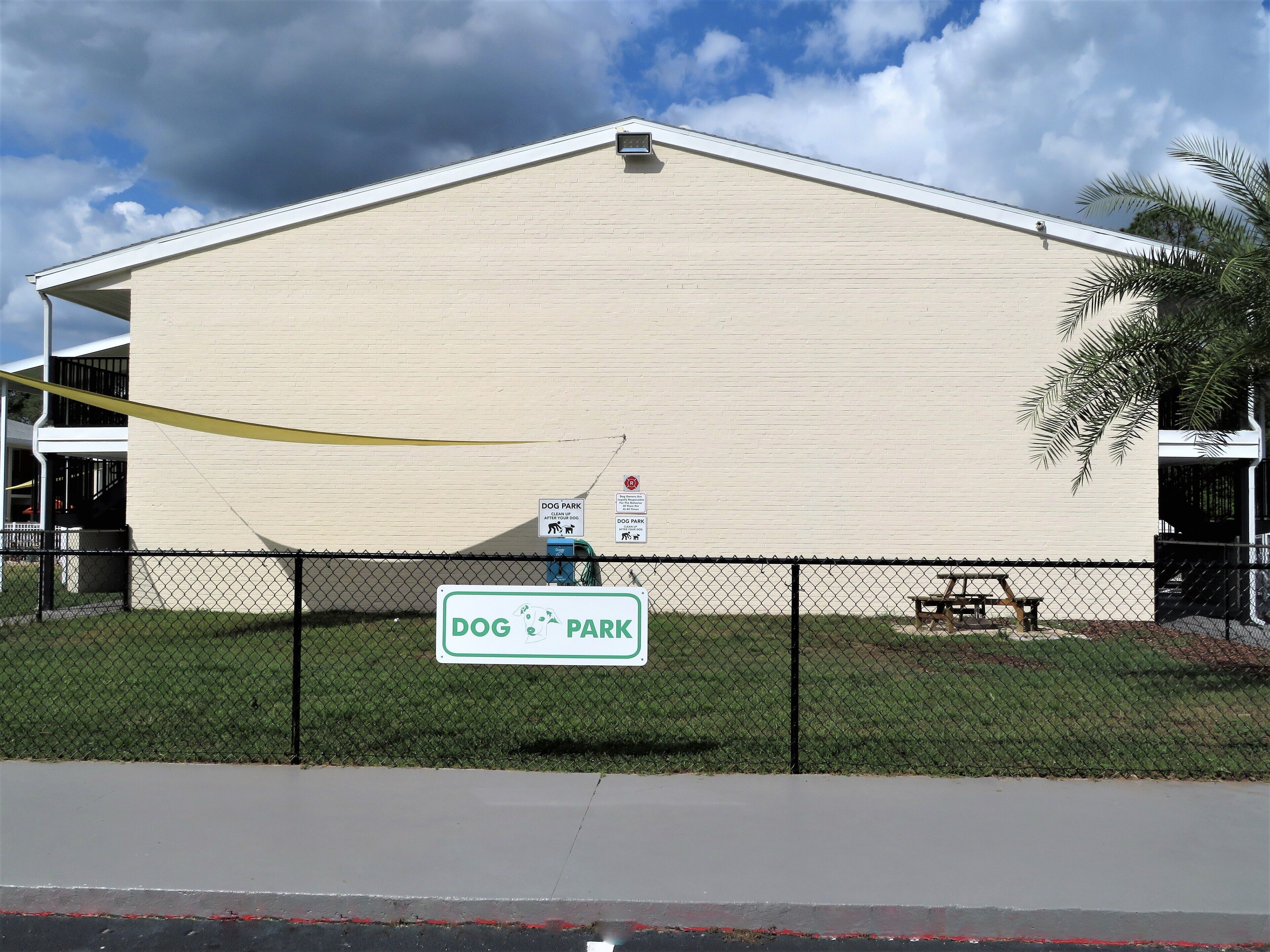 Un bâtiment sur lequel on peut lire l’inscription « PARC POUR CHIENS », une zone délimitée par une clôture et un banc.