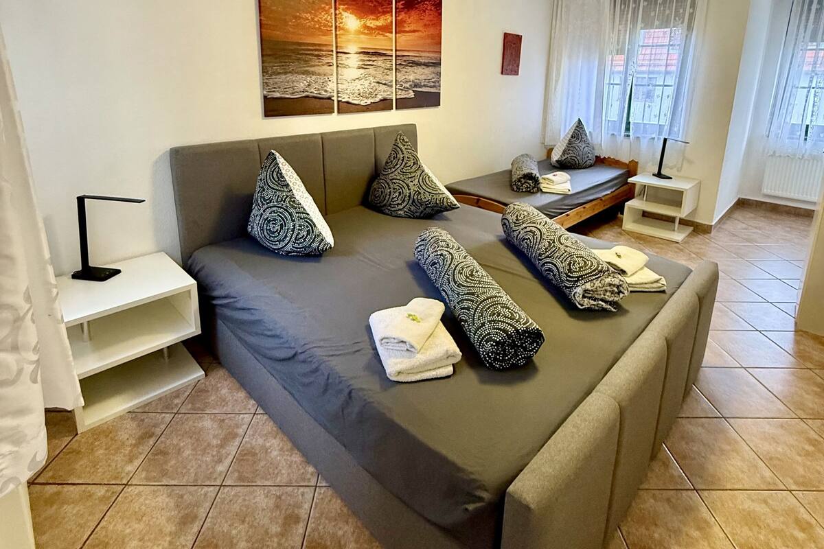 A bedroom with a large bed, a bedside table, a nightstand, a window with curtains, and a wall-mounted artwork.