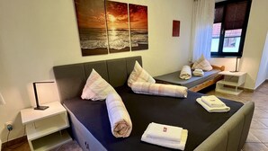 Cama bem arrumada com toalhas, mesa de cabeceira, abajur e um quadro de um pôr do sol sobre o oceano na parede.
