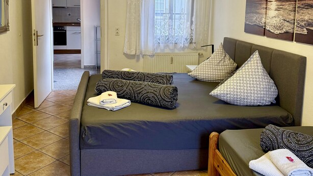 Quarto com cama, janela com cortinas, quadro na parede e vista para a cozinha através de uma porta aberta.