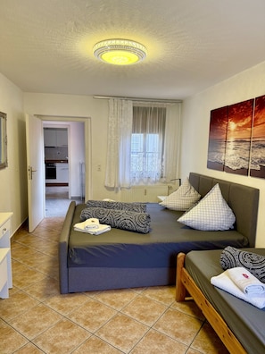 A bedroom with a bed, a window with curtains, a painting on the wall, and a view of the kitchen through an open door. - Appartements Rehn (Dresden)