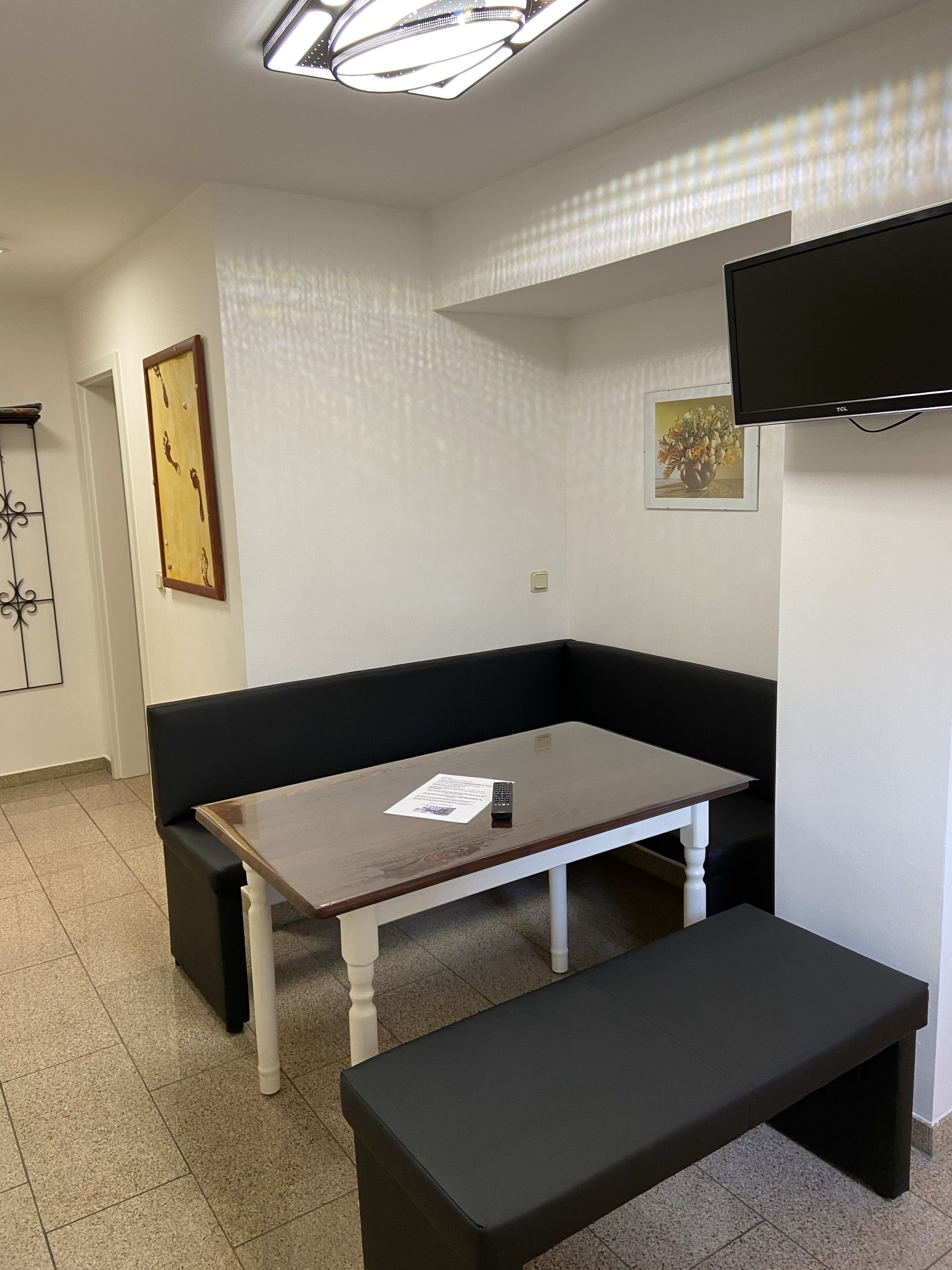 A room with a black bench, a wooden table, a black sofa, a flat-screen TV mounted on the wall, and a framed picture on the wall.