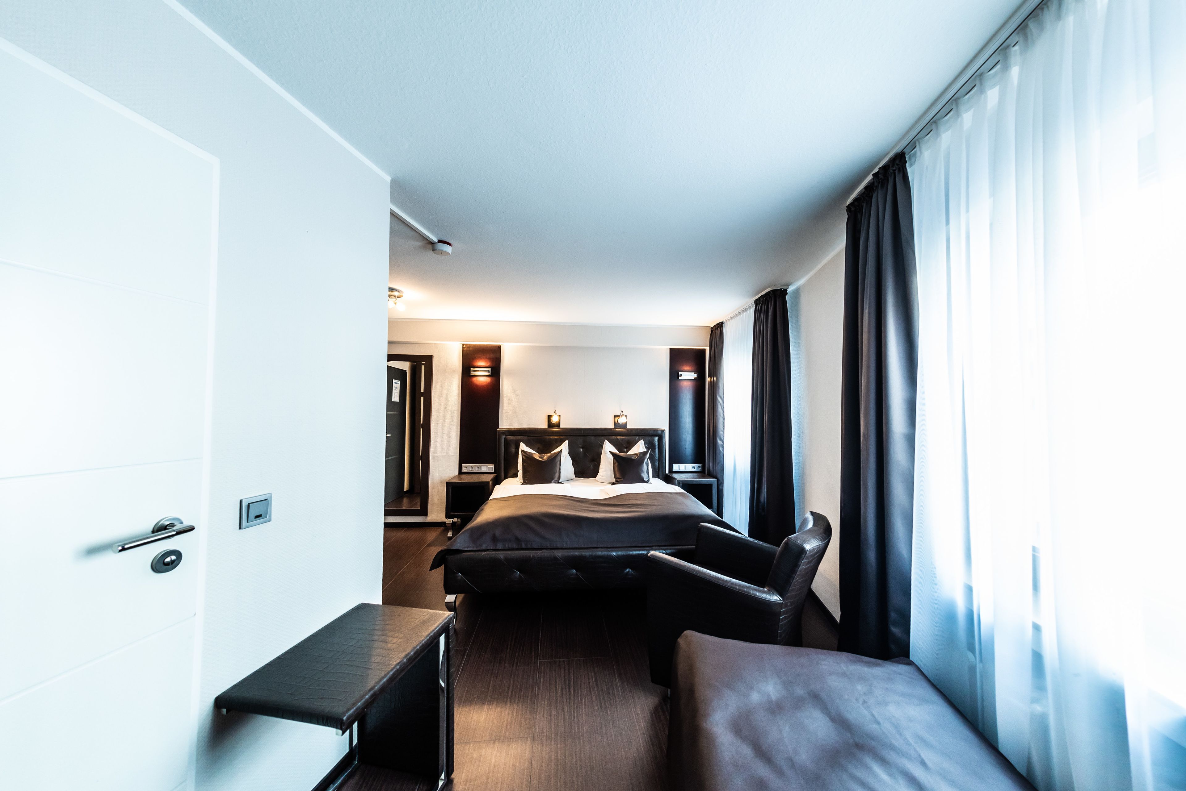 A modern hotel room with a bed, a desk, and a chair. There is a door and a window with curtains.