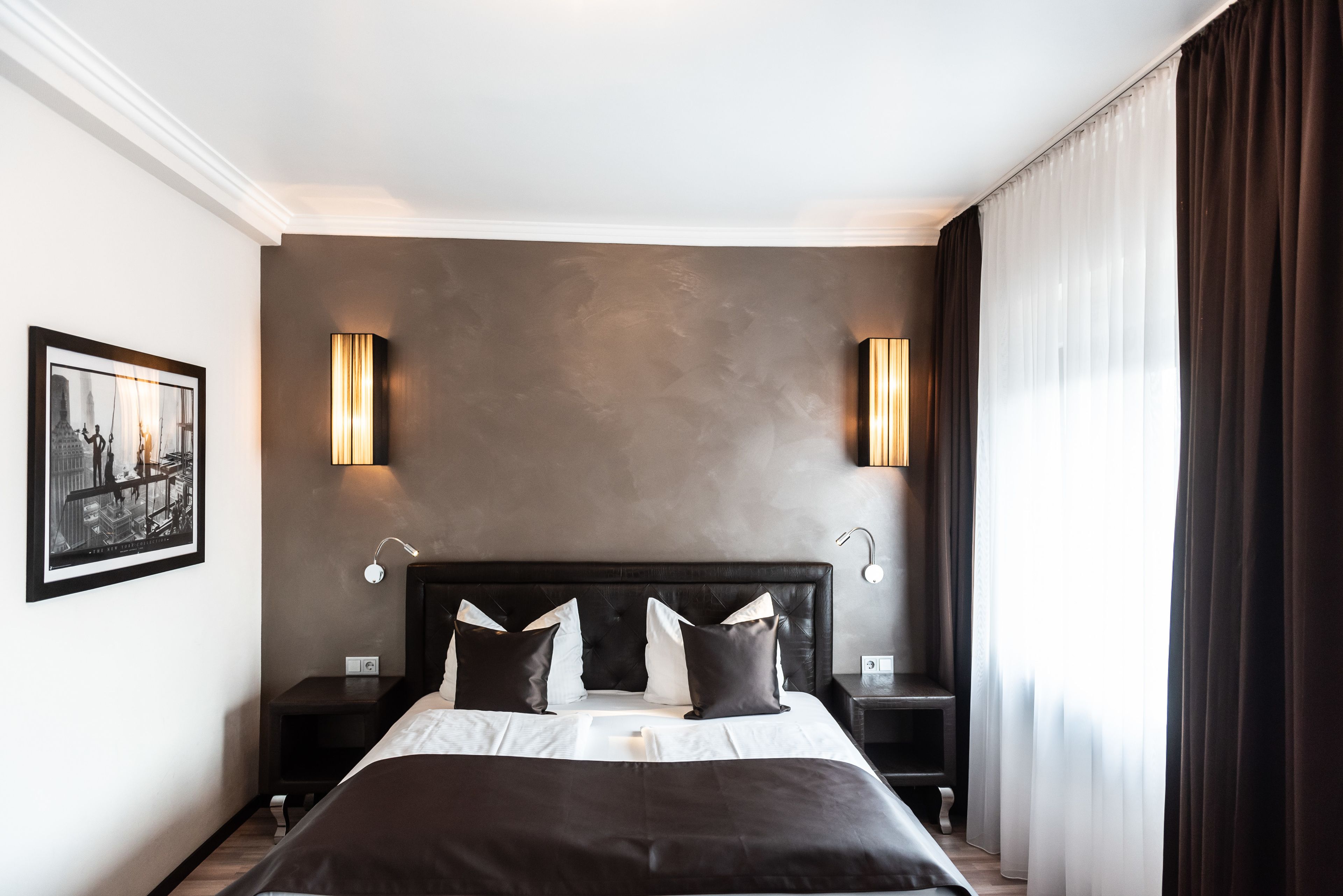 A modern hotel room with a large bed, two bedside tables, a wall-mounted lamp, and a framed picture on the wall.