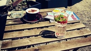 Una mesa de madera con una bebida, un plato de comida y una revista. Hay plantas en macetas negras y un edificio al fondo.