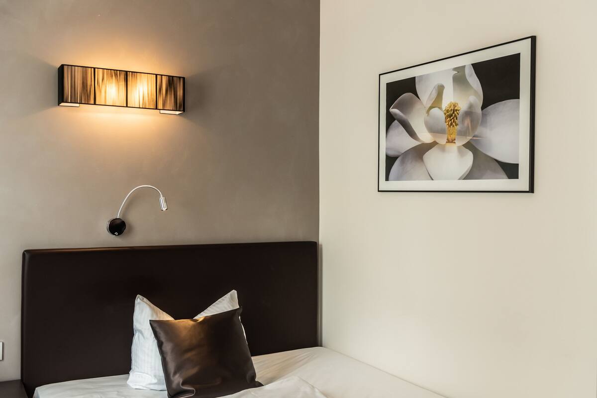 Un lit bien fait, avec une tête de lit sombre, un luminaire fixé au mur et un tableau représentant une fleur.