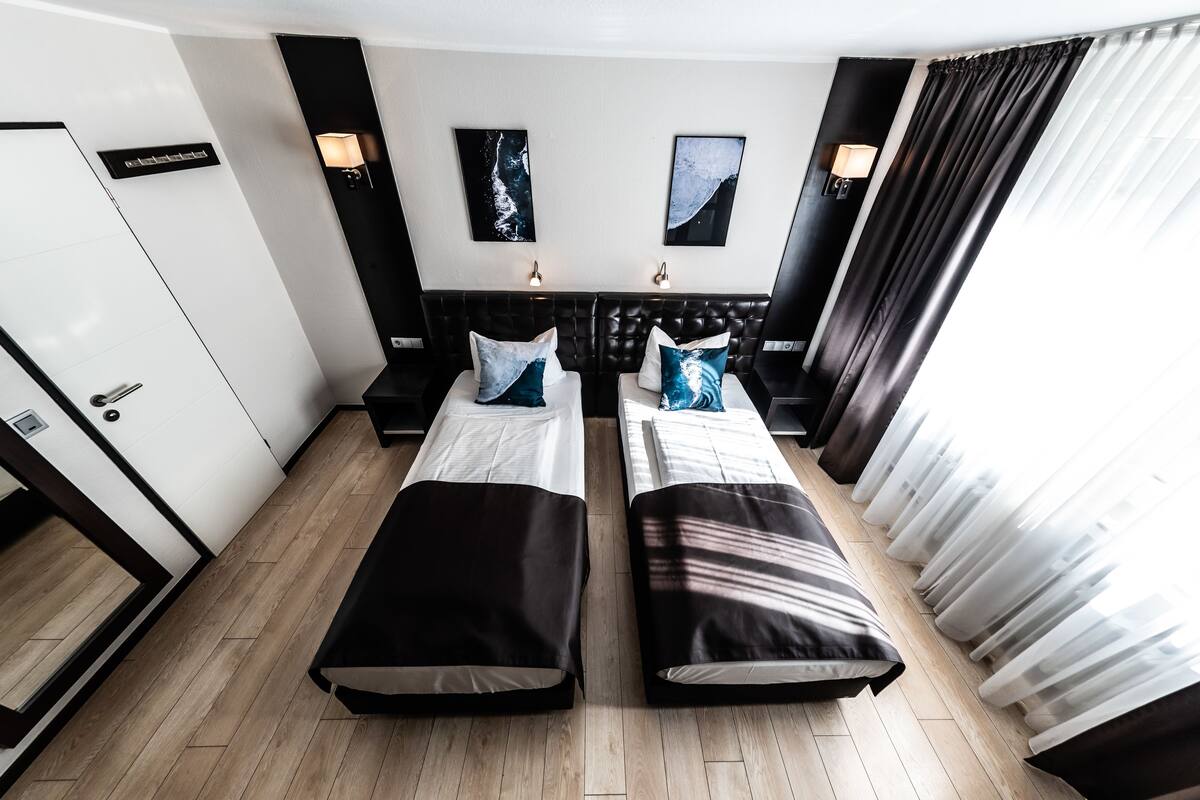 Une chambre d’hôtel avec deux lits simples, une tête de lit sombre, deux lampes fixées au mur et une grande fenêtre ornée de rideaux légers.