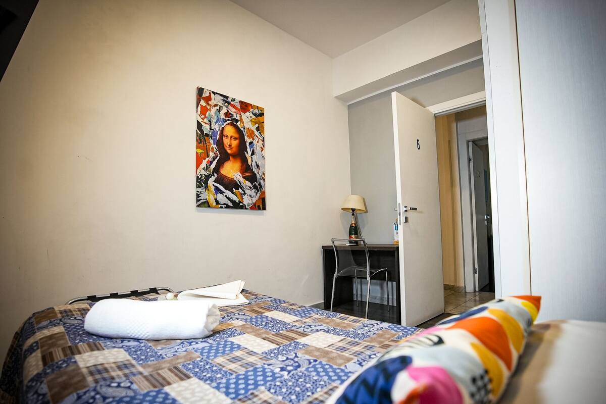 A hotel room with a bed, a desk, a chair, a painting on the wall, and a door leading to another room.