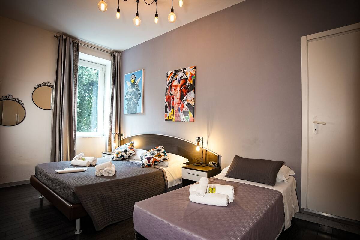 A hotel room with two beds, a large window, a painting, and a bedside table with a lamp.