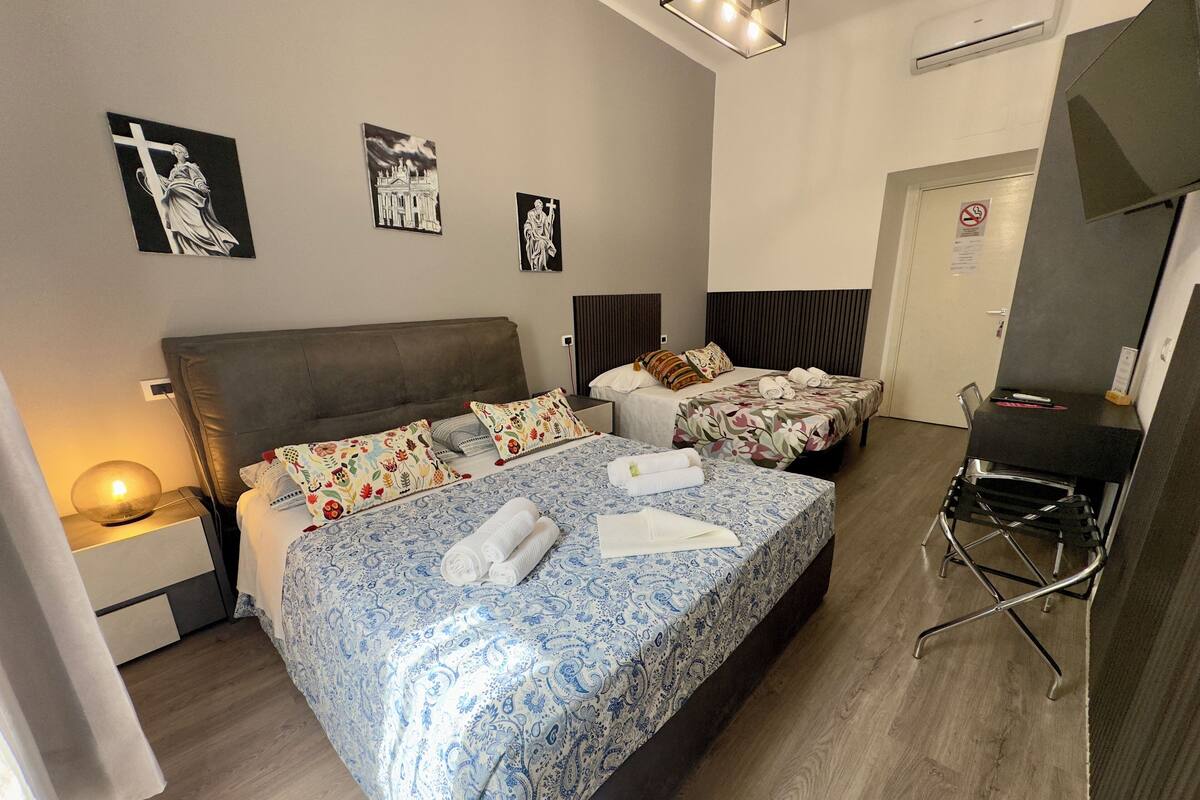 A hotel room with a large bed, two smaller beds, a desk, and a chair. There are wall art pieces and a lamp on the nightstand.