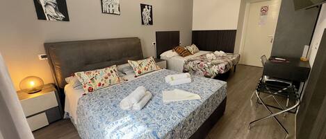 Habitación de hotel con una cama grande, dos camas más pequeñas, un escritorio y una silla. Hay obras de arte en la pared y una lámpara en la mesita de noche.