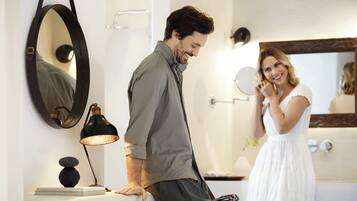 A man leaning on a counter with a lamp and books beside him, and a woman in a white dress standing by a mirror.