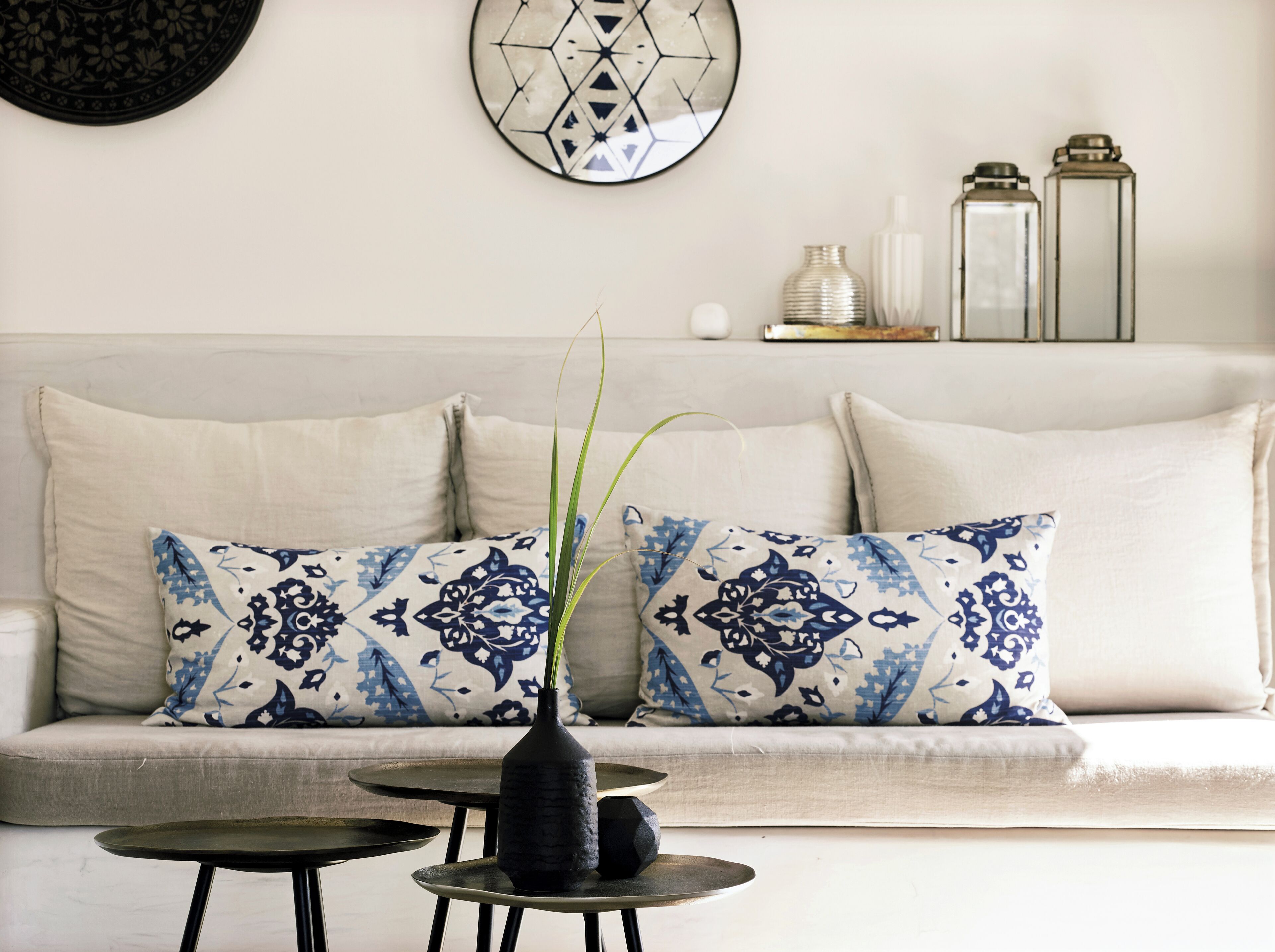 A modern living room with a sofa, patterned cushions, a vase with a plant, and a decorative wall piece.