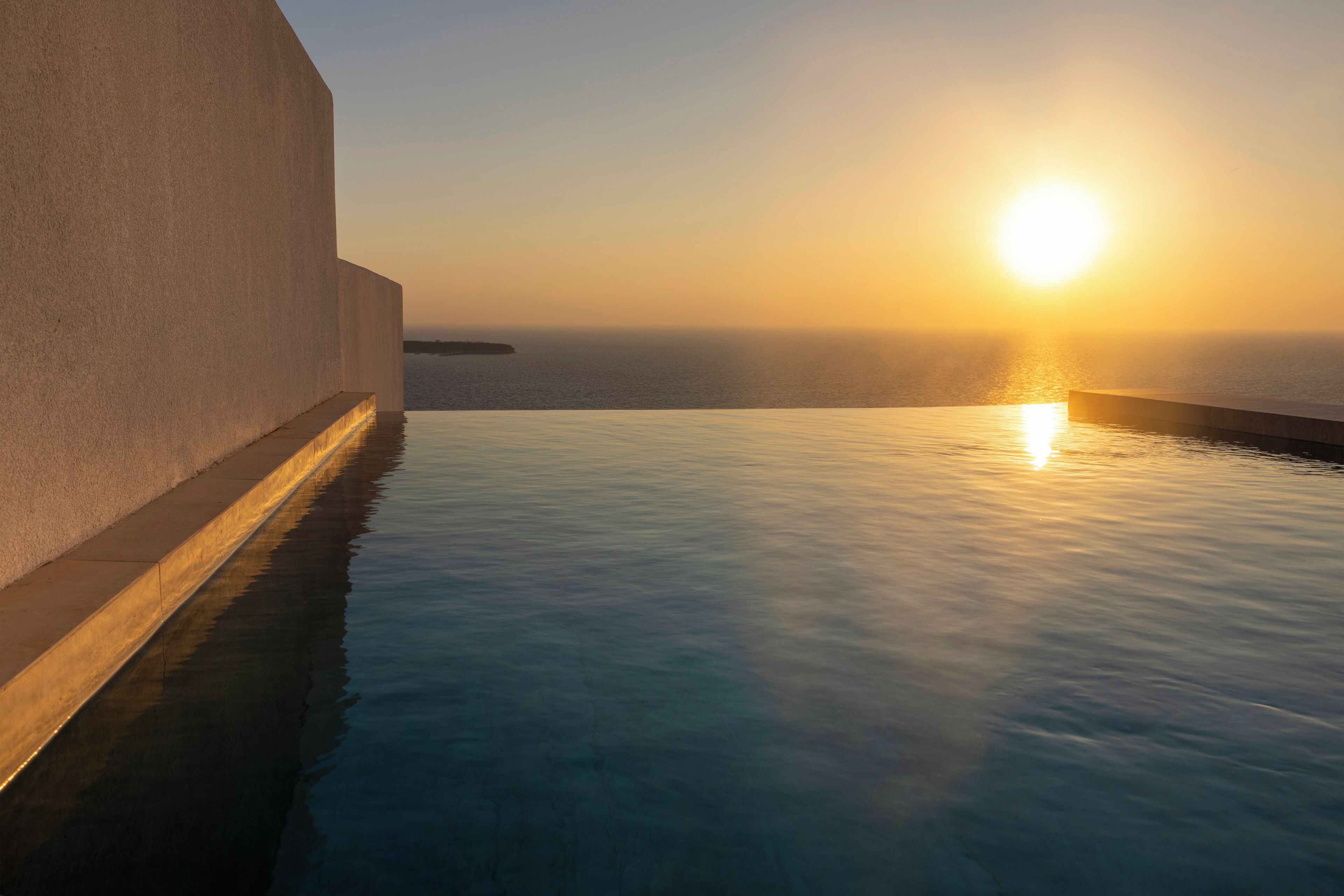 A serene infinity pool with a sunset view, a smooth white wall, and calm water reflecting the sky.