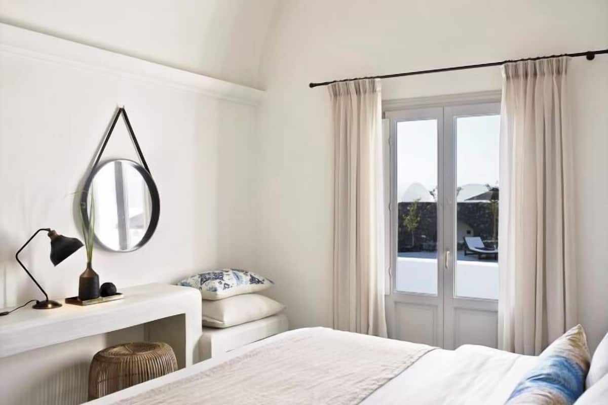 Ein Schlafzimmer mit Bett, Nachttisch, Lampe, Spiegel und einem Fenster mit Vorhängen.