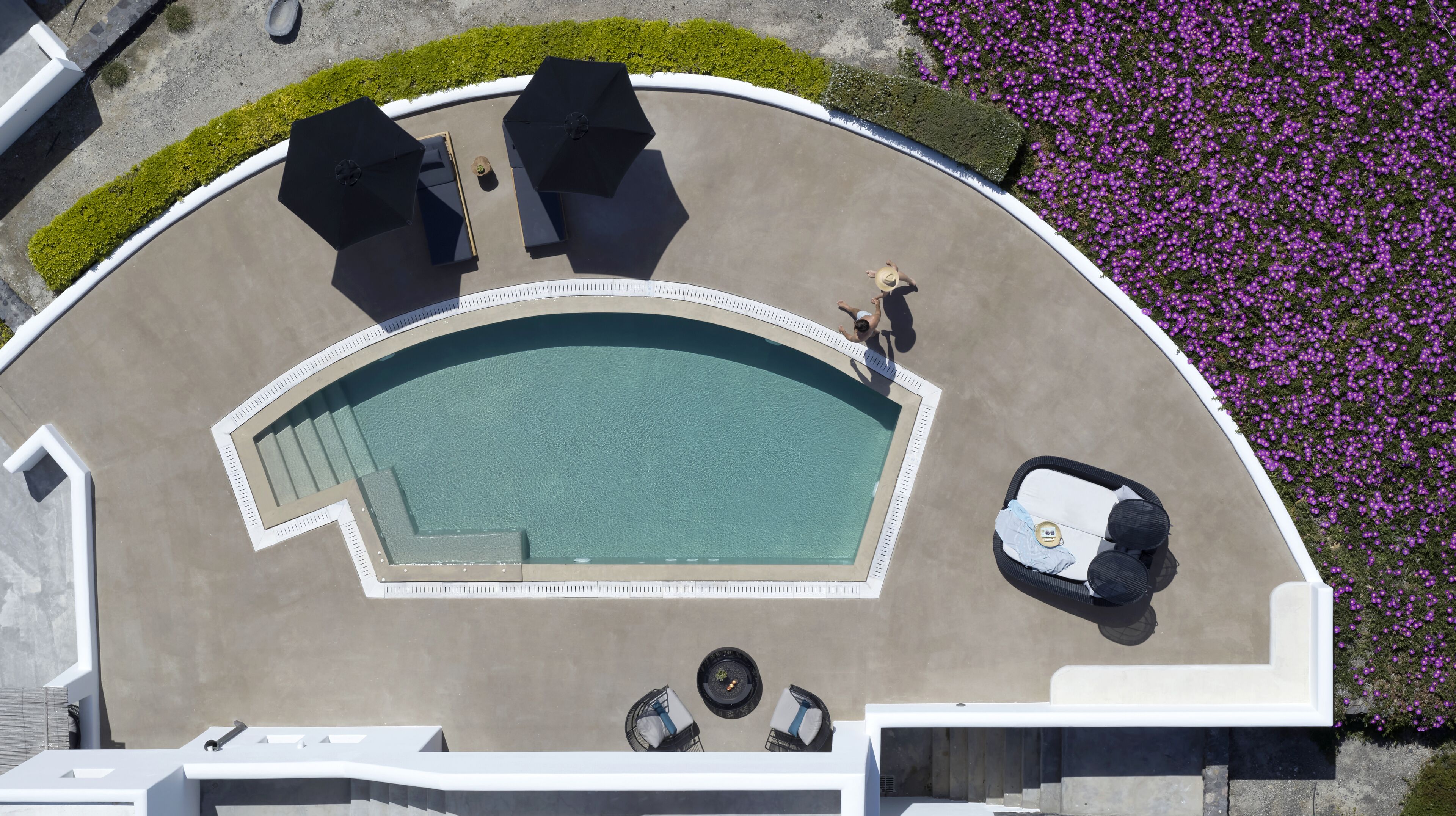 An aerial view of a pool area with a curved pool, two umbrellas, a small table with chairs, and a car parked nearby.