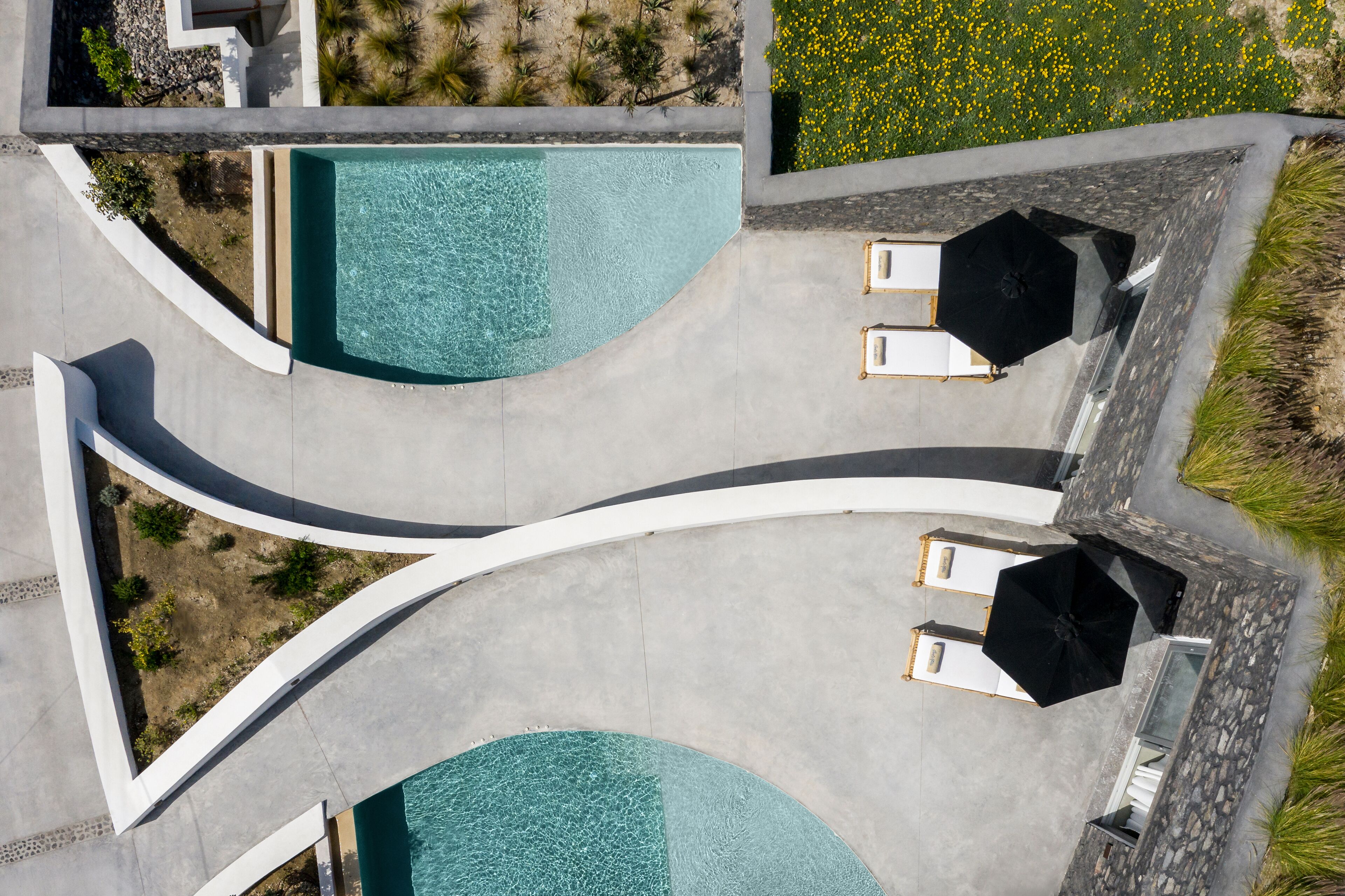 Vista aérea de un área de piscina con dos sillones de descanso y sombrillas, rodeada por un jardín ajardinado con flores amarillas.