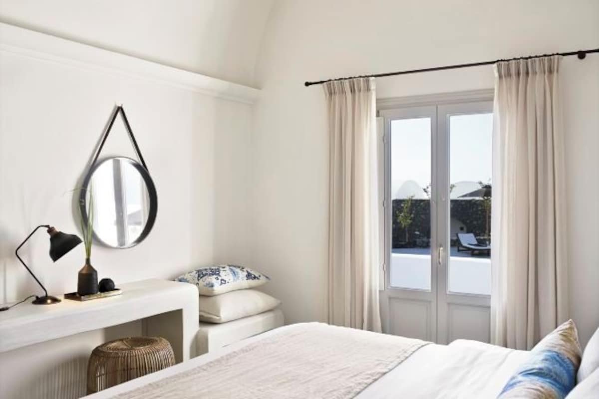 A bedroom with a bed, a bedside table, a lamp, a mirror, and a window with curtains.
